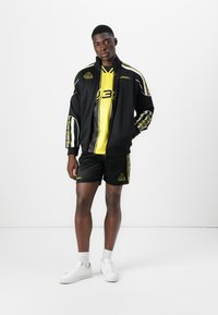Black sporty jacket with yellow accents, matching yellow shirt, black shorts, and white sneakers with textured soles.