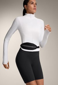 Bela dolga sleeve zip-up jakna, oblečena čez črne oprijete kratke hlače, z črno torbico z belimi poudarki, gladko tkanino in športno prileganje.