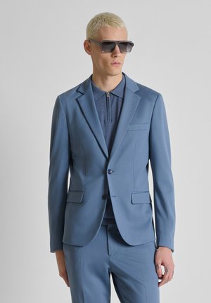 Uomo biondo con occhiali da sole scuri, indossa una camicia blu con zip, una giacca e pantaloni abbinati blu su uno sfondo semplice.