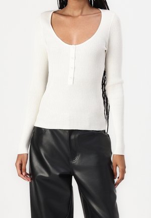 Kobieta ubrana w biały prążkowany, długi rękaw, zapinany na guziki top z dekoltem typu scoop oraz czarne, skórzane spodnie z wysokim stanem.