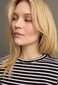 Gestreiftes Schwarz-Weiß-Hemd mit Rundhalsausschnitt, kombiniert mit einer filigranen goldenen Kettenhalskette. Blonde Haare fallen sanft um das Gesicht.