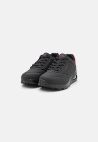 Black athletic shoes with textured overlay, round toe, lace-up design, and pink accents on the tongue and heel, featuring a cushioned sole.