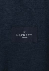 Tmavomodrá tkanina s prišitým obdĺžnikovým štítkom nesúcim nápis "Hackett London" a malým logom so skríženými tenisovými raketami nad textom.