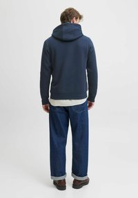 Sudadera azul marino con capucha amplia, confeccionada en una tela suave, llevada sobre una camisa blanca, combinada con jeans de corte holgado y zapatos marrones.