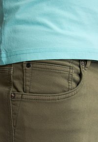 T-shirt en coton turquoise superposé sur un pantalon vert olive, affichant une poche avant avec des détails de couture et un accent en métal.