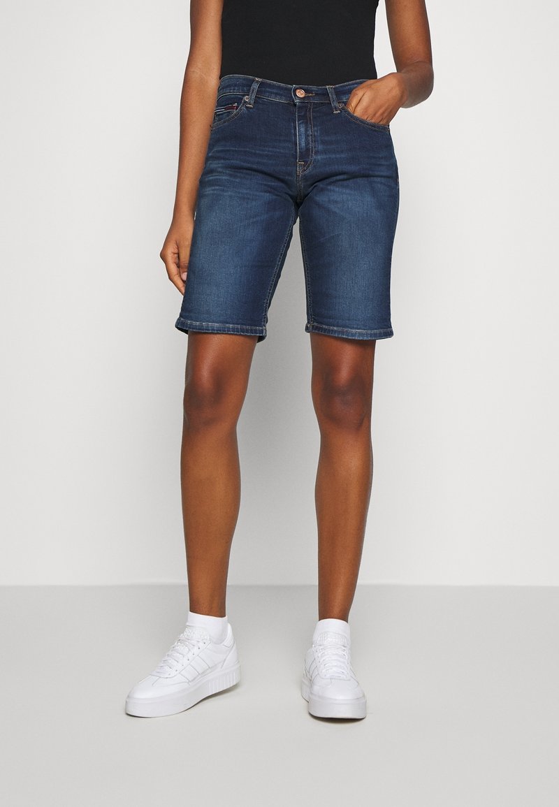 Pantalones cortos de mezclilla en azul oscuro con un diseño ajustado, cinco bolsillos y dobladillo enrollado. Combinados con zapatillas blancas.