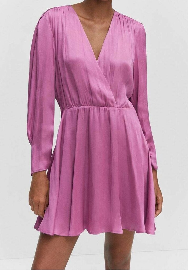 Femme portant une robe rose à manches longues avec un profond décolleté en V et une taille froncée, debout devant un fond clair uni.