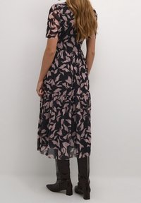 Maxikleid mit navyblauem Grund, das ein pinkes Blättermuster aufweist, kurze Ärmel und einen gestuften Rock hat. Getragen mit knielangen schwarzen Stiefeln mit Krokodilstruktur.