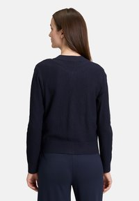 Navyfarbener strukturierter Cardigan mit langen Ärmeln und geripptem Saum, runder Halslinie und lässigem Schnitt. Getragen über navyfarbenen Hosen.