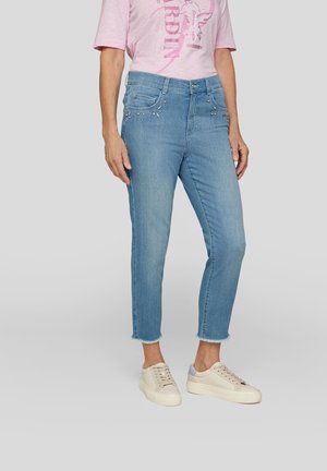 Light blue denim jeans with a straight cut, frayed hem, and decorative embellishments on the front pockets, paired with beige sneakers.