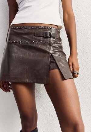 Femme portant une mini-jupe en cuir marron avec des clous métalliques et une boucle, associée à un haut court blanc, debout devant un mur texturé blanc.