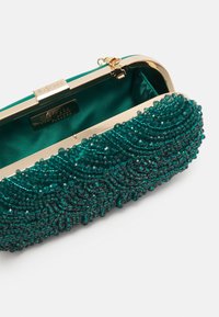 Kopertówka w kolorze morskim, ozdobiona koralikami, z złotą ramą, z misternymi falistymi wzorami i satynową podszewką. Wygląda elegancko i kompaktowo.