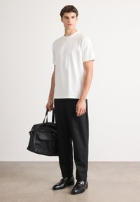 T-shirt en coton blanc à col rond, associé à un pantalon noir ajusté et des chaussures en cuir noir. Le modèle tient un sac en cuir noir.