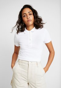 Fehér, slim fit pólóing, gallérral, rövid ujjal �és gombos kivágással, bézs, magas derekú nadrággal párosítva.