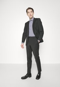 Completo nero con giacca aderente sopra una camicia a righe blu e bianche. Pantaloni slim fit e scarpe nere lucide completano il look.