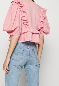 Růžová blůzka s volány, krátkými nabíranými rukávy, vrstveným lemem a lehkým materiálem, v kombinaci se světle modrými džíny.