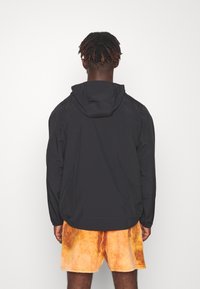 Man seen from behind wearing a black hooded jacket and orange tie-dye shorts standing against a plain white background.