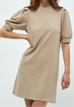 Beige jurk met korte mouwen en geplooide mouwen met geribbelde manchetten, gedragen door een persoon met een hand op de heup.