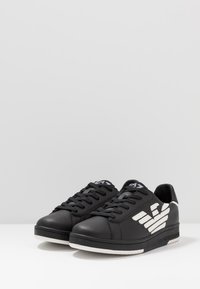 Baskets en cuir noir avec des accents de logo blancs, bout rond, semelle plate et design classique à lacets ; surface texturée avec une finition en cuir lisse.