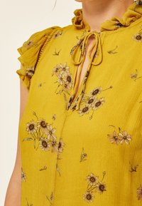 Blouse florale jaune avec manches courtes à volants et encolure avec ouverture en goutte d'eau nouée, portée par une personne.
