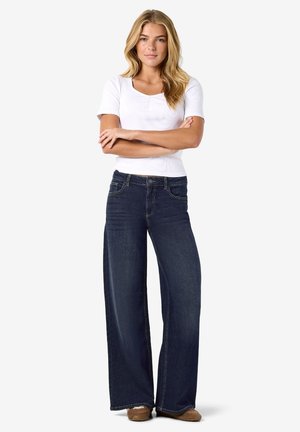 Femme aux longs cheveux blonds, debout, les bras croisés, portant un haut blanc à manches courtes, un jean bleu foncé à jambes larges et des chaussures marron.