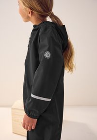 Black waterproof jacket with a hood, featuring elastic cuffs, a reflective stripe, and a circular logo patch on the left sleeve.