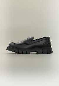 Mocassin noir à semelle épaisse, dessus en cuir lisse avec détail de bride, présenté de côté sur un fond neutre.