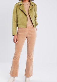 Veste de motard en daim vert olive avec fermeture éclair et accents noirs, associée à un haut côtelé marron clair et à un pantalon en velours côtelé évasé.