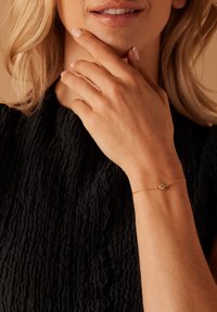 Gouden kettingarmband met een kleine, ronde edelsteen. Gewrongen zwart stoffen achtergrond. Hand rust nabij de kin en toont de armband.