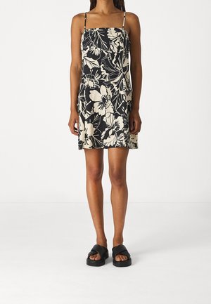 Vrouw draagt een mouwloze mini-jurk met zwart-witte bloemenprint en dunne bandjes, gecombineerd met zwarte open-toe slides.