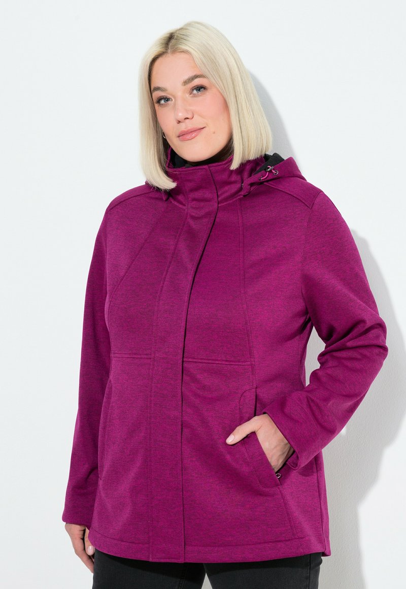 Kobieta z krótkimi blond włosami ma na sobie fioletową kurtkę z kapturem, trzyma jedną rękę w kieszeni z przodu, stojąc na tle białym.