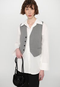 Gilet grigio in lana dal taglio sartoriale, con bottoni neri e due tasche laterali, indossato su una camicia bianca. Tiene una piccola borsa nera.