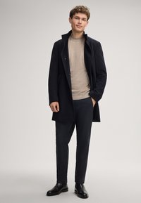 Dunkler Wollmantel, beiger Strickpullover und dunkle strukturierten Hosen. Glatte schwarze Stiefel runden das Outfit ab, alles vor einem neutralen Hintergrund.