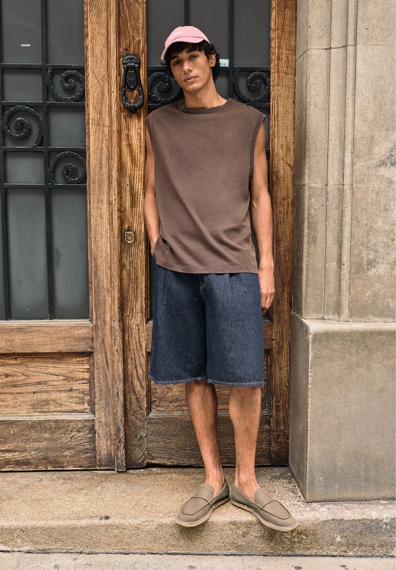 Jeune homme portant un débardeur marron, un short en jean, une casquette rose et des mocassins beiges, debout devant une porte en bois avec des grilles métalliques noires décoratives.