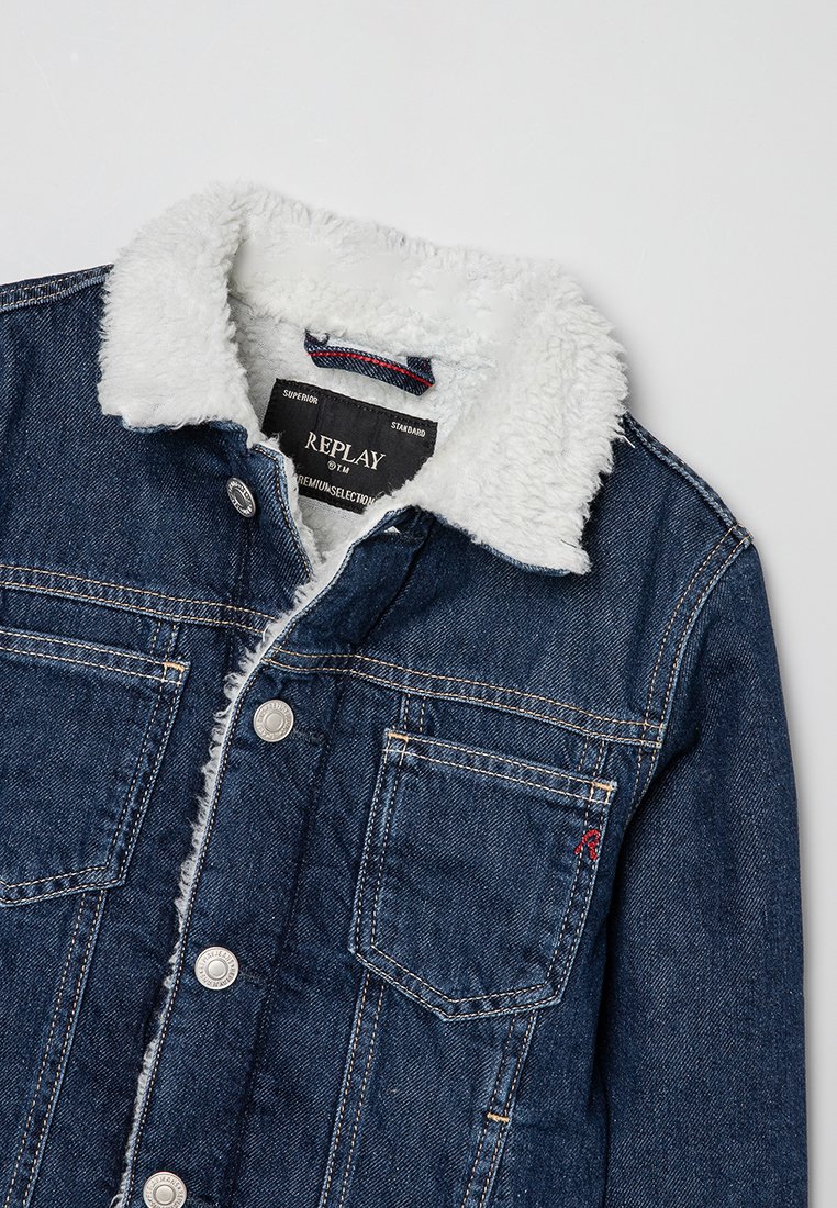 Chaqueta de mezclilla azul con cuello y forro de felpa blanca, cierre de botones, bolsillos en el pecho y etiqueta de la marca Replay en el interior del cuello.