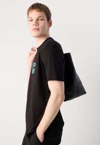Junger Mann in schwarzem T-Shirt, der eine schwarze Ledertasche über der Schulter trägt, steht vor einem schlichten hellen Hintergrund.