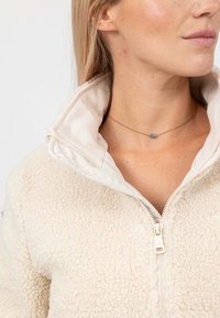 Giacca in pile color crema con un esterno testurizzato e un morbido rivestimento interno, dotata di chiusura a zip e collo alto. Include una semplice collana d'oro.