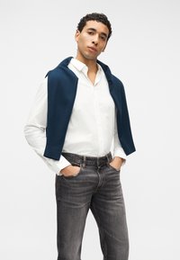 Νεαρός άνδρας με λευκό πουκάμισο με κουμπιά, σκούρο γκρι τζιν, με μπλε ναυτικό πουλόβερ ριγμένο στους ώμους, χέρια στις τσέπες, ουδέτερο φόντο.