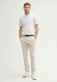 Jeune homme debout avec les mains dans les poches, portant un t-shirt blanc, un pantalon beige, une ceinture noire et des baskets blanches sur fond uni.