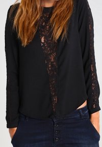 Blouse noire avec des manches longues et des détails en dentelle le long du centre et des manches ; tissu doux avec une silhouette légèrement drapée.