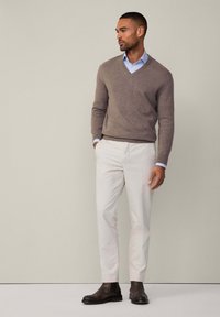 Mies, joka on pukeutunut ruskeaan V-kaula-aukkoiseen villapaitaan, vaaleansiniseen paitaan, valkoisiin housuihin ja tummanruskeisiin nahkasaappaisiin, seisoo yksinkertaista beigeä taustaa vasten.