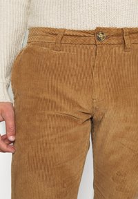 Bruine broek van corduroy met verticale ribbels, voorzien van een knoopsluiting en riemlussen, getoond naast een crèmekleurige trui met textuur.