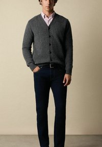 Šedý vlněný cardigan s výstřihem do V, vyznačující se tmavými knoflíky. Kombinovaný s tmavě modrými džíny a růžovou košilí s límečkem. Jednoduchý, texturovaný design.