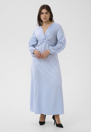 Frau trägt ein hellblaues Maxikleid mit langen Ärmeln, einer gedrehten Vorderteil-Taillendetail und schwarzen High Heels, steht vor einem schlichten Hintergrund.