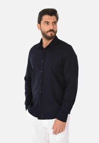 Camicia a maniche lunghe di colore blu navy realizzata in tessuto morbido, con colletto button-down, bottoni frontali e polsini bottonati; indossata con pantaloni bianchi.