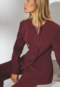 Vyno raudonos spalvos pritaikytas bleizeris su dvigubais mygtukais, plačiomis rankovėmis ir elegantiška siluetu. Sklandus audinys su subtiliu blizgesiu.