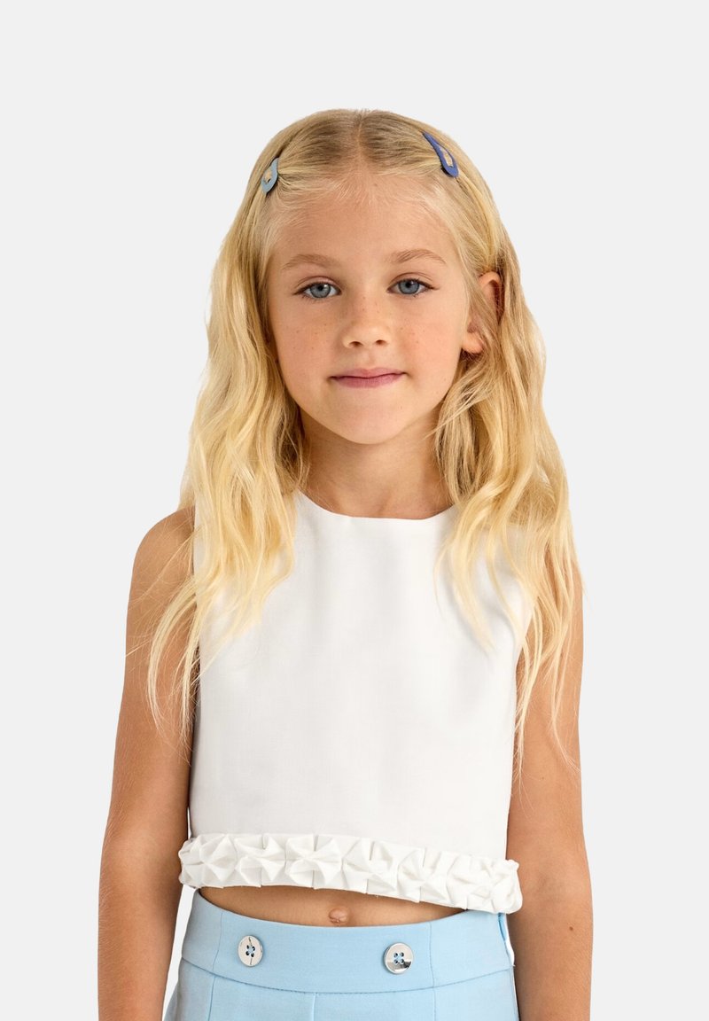 Top bianco corto con orlo a volant testurizzato, abbinato a pantaloni azzurri chiari a vita alta con bottoni decorativi. Tessuto morbido, design senza maniche.