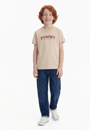 Smilende rødhåret dreng iført beige Tommy Hilfiger t-shirt, blå jeans og hvide sneakers, stående mod en hvid baggrund.
