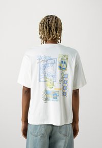Camiseta blanca de algodón con impresiones gráficas azules de mapas y arquitectura de Lisboa, con acentos de hojas verdes y detalles estampados.
