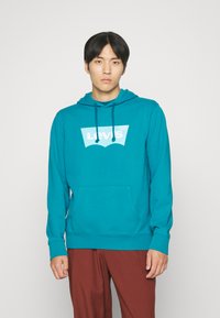 Teal hoodie met trekkoorden, groot wit Levi's-logo, voorkant zak, ribgebreide manchetten en zoom. Model gecombineerd met bruine broek.